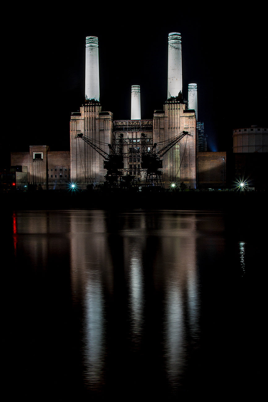 The iconic Battersea Power Station with it's 4 tall white towers illuminated at night and reflected on the Thames with two cranes at the waters edge.