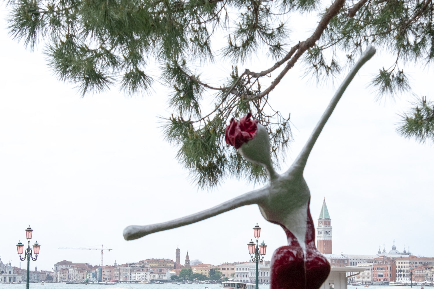 A sculpture of a woman in a red backless dress with outstretched arms reaching for the sky and the Venice skyline at the Venice Biennale 2019. 