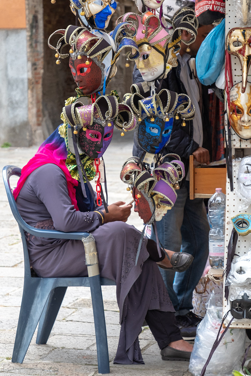 The juxtaposition of a preoccupied stall holder looking at her phone with her face hidden behind one of her many Venetian masks. 