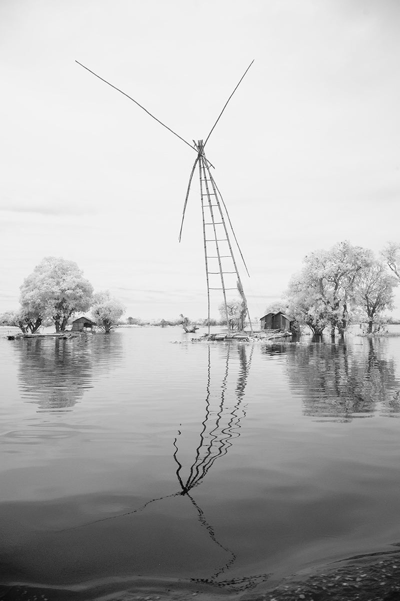 A monochrome image of a fishing frame and trees on the Sangker river taken from the boat from Siem Reap to Battambang in Cambodia. Nets are attached to the fishing frame to catch the fish.