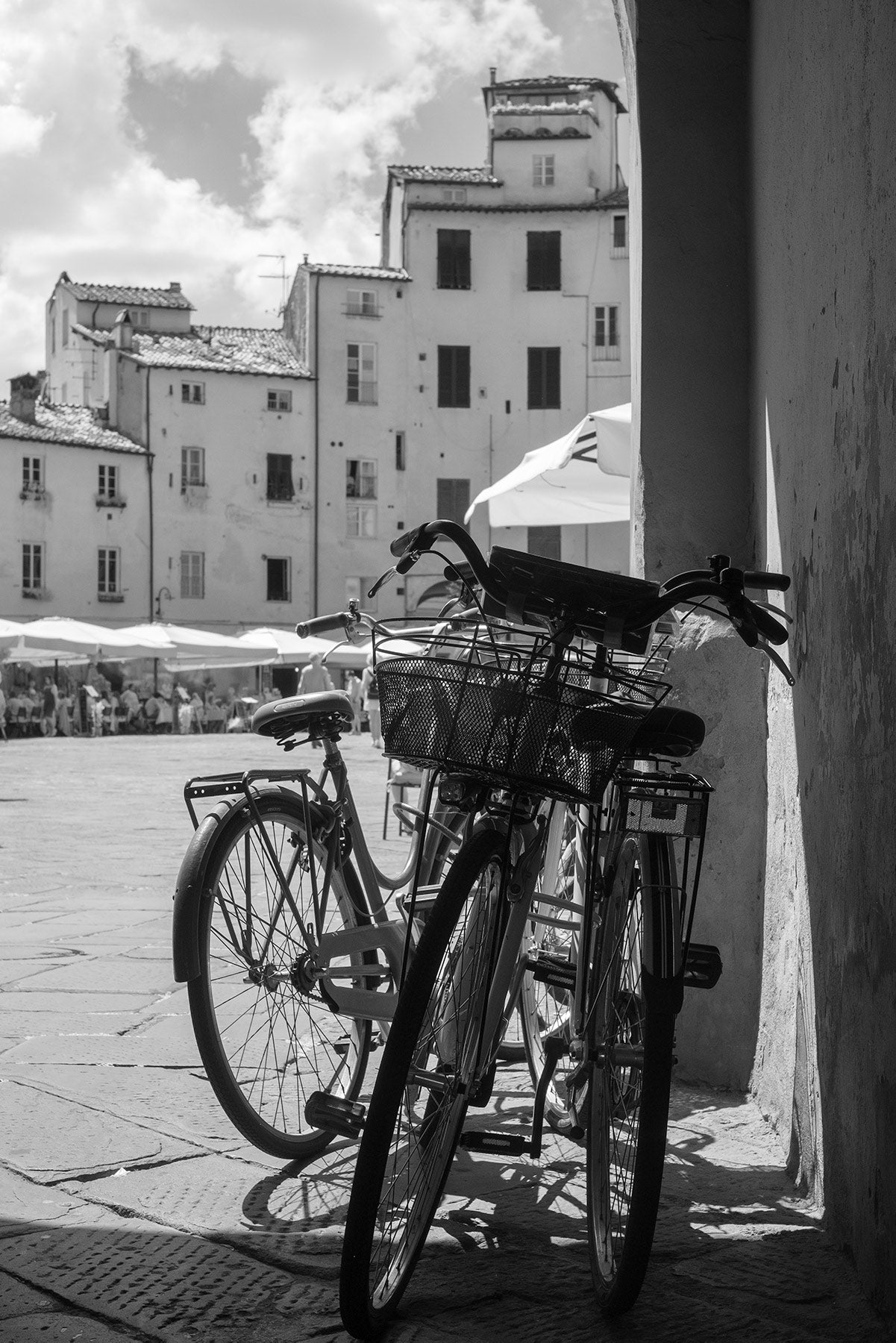 Black and White image looking past three bicycles leant against a wall into Piazza dell'Anfiteatro, the oval shaped piazza in Lucca, Italy.